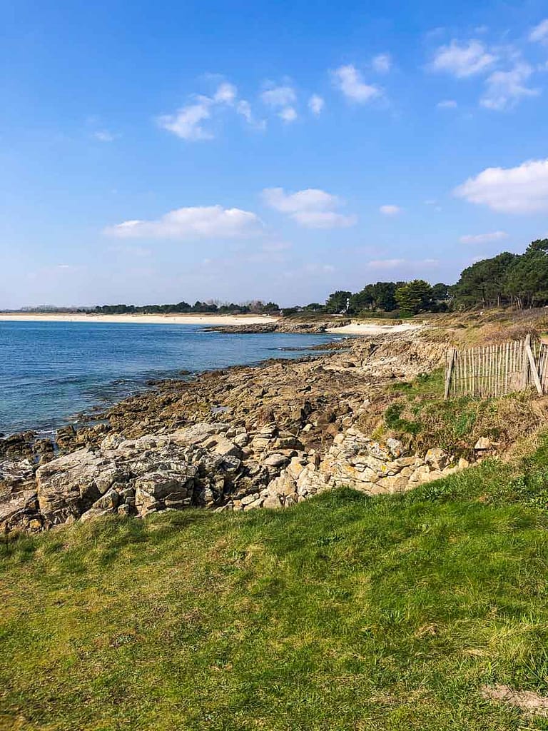 The rocky shoreline and grassy area, encircled by a picturesque fence, unfold under a blue sky dotted with clouds, creating an incomparable atmosphere reminiscent of the serene port of Sainte Marine.