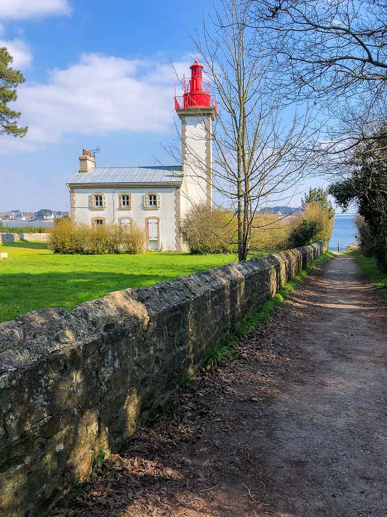 A stone path leads to a white lighthouse with a red top, surrounded by greenery and trees under a blue sky, capturing the incomparable atmosphere of the charming port of Sainte Marine.